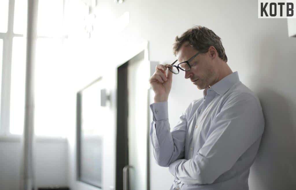 Thoughtful man in a bright room holding his glasses while leaning against a wall.